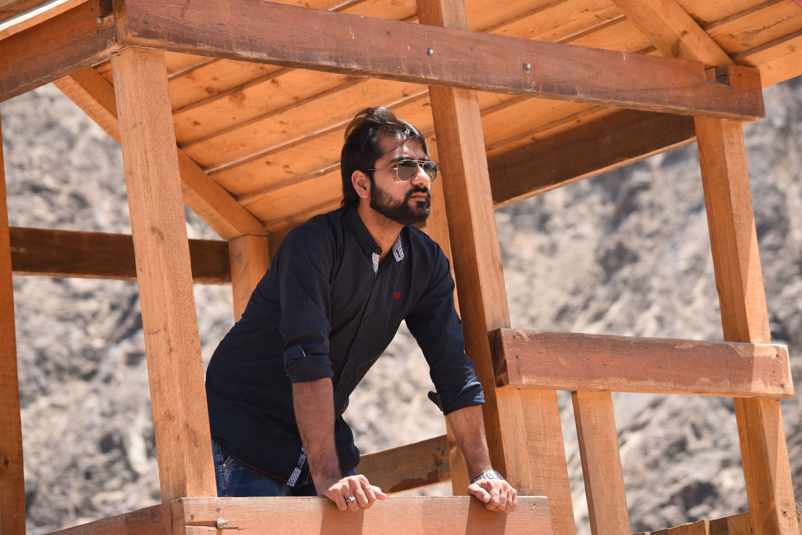 A man with sunglasses leans on a wooden structure outdoors, under clear daylight.
